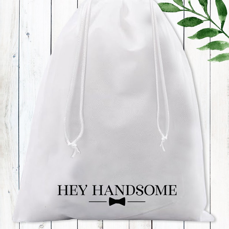 Puede incluir: Bolsa de tela blanca con cord&oacute;n de ajuste, con el texto "HEY HANDSOME" y un lazo negro.