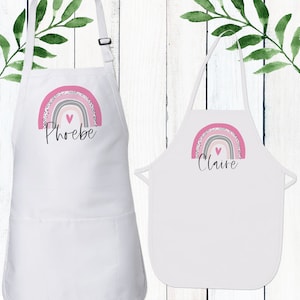 May include: Two white aprons with pink and grey rainbow designs and a pink heart. The aprons have the names "Phoebe" and "Claire" printed on them.