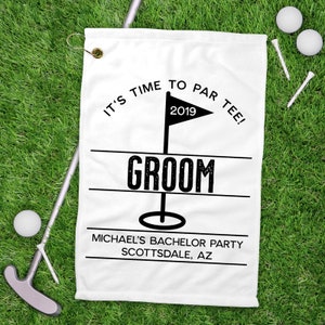 May include: White golf towel with the text "IT'S TIME TO PAR TEE!" and "GROOM". Includes a flag with the year 2019. The towel also has the text "MICHAEL'S BACHELOR PARTY SCOTTSDALE, AZ". Golf balls, tees, and a putter are on the green grass.