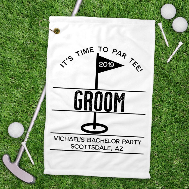 May include: White golf towel with the text "IT'S TIME TO PAR TEE!" and "GROOM". Includes a flag with the year 2019. The towel also has the text "MICHAEL'S BACHELOR PARTY SCOTTSDALE, AZ". Golf balls, tees, and a putter are on the green grass.