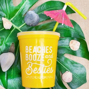 May include: Yellow plastic cup with the text "Beaches Booze and Besties" printed on it. A green leaf with a pink and white umbrella and a yellow straw is in the background.