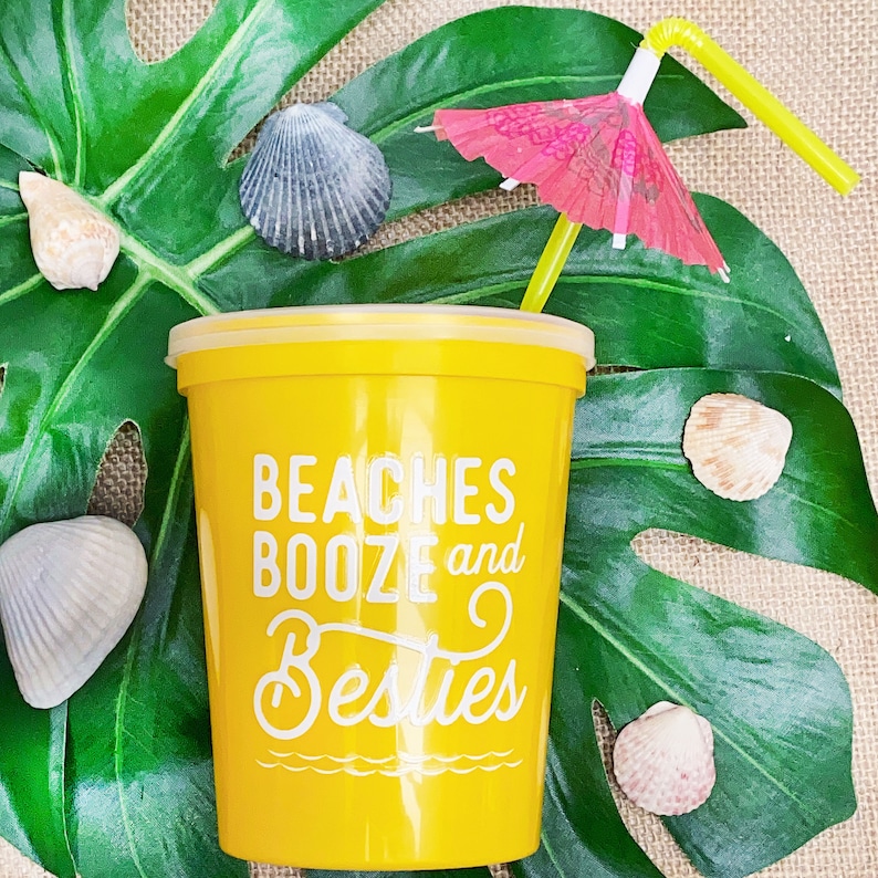 May include: Yellow plastic cup with the text "Beaches Booze and Besties" printed on it. A green leaf with a pink and white umbrella and a yellow straw is in the background.