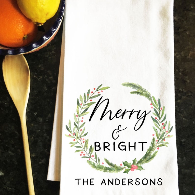 May include: White kitchen towel with a green wreath design and the text "Merry & Bright The Andersons".