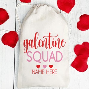 May include: White canvas bag with a red "galentine squad" text and pink "NAME HERE" text. Three pink hearts are below the text. The bag is surrounded by red heart-shaped petals.