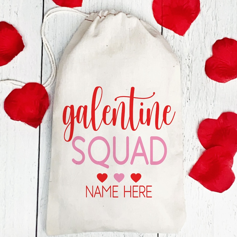 May include: White canvas bag with a red "galentine squad" text and pink "NAME HERE" text. Three pink hearts are below the text. The bag is surrounded by red heart-shaped petals.