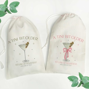 May include: Two white cotton drawstring bags with the text "A TINI BIT OLDER" printed on them. Each bag features a martini glass with olives and a pink ribbon. The text "1ST LINE OF YOUR TEXT" and "2ND LINE OF TEXT" is printed below the martini glass.
