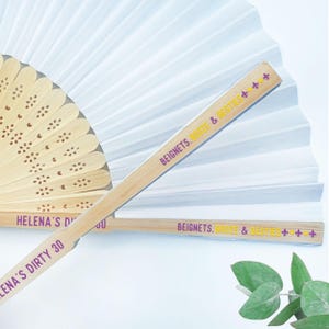 May include: A close-up shot of two bamboo fans with white paper fan backgrounds. One fan is open, and the other is closed. The text "BEIGNETS, BOOZE & BESTIES" is printed on the fan handle. The text "HELENA'S DIRTY 30" is printed on the other fan handle.