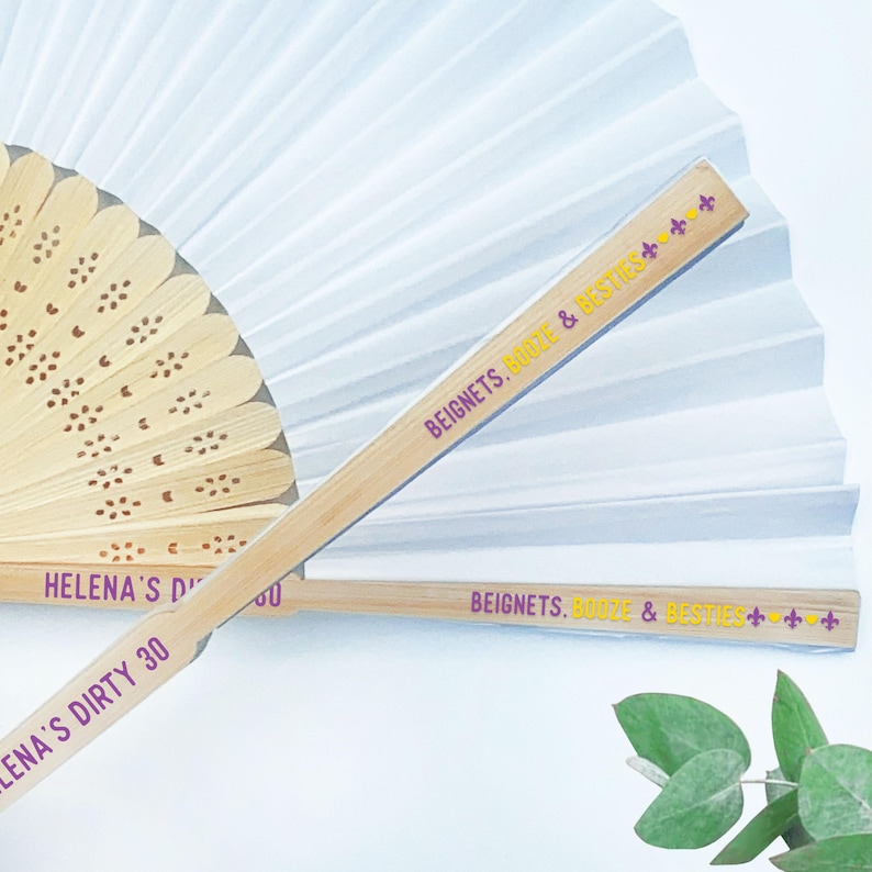 May include: A close-up shot of two bamboo fans with white paper fan backgrounds. One fan is open, and the other is closed. The text "BEIGNETS, BOOZE & BESTIES" is printed on the fan handle. The text "HELENA'S DIRTY 30" is printed on the other fan handle.