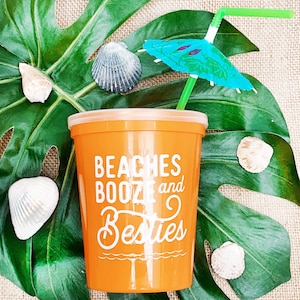 May include: Orange plastic cup with the text "Beaches Booze and Besties" printed on it. A green straw with a blue and green umbrella is sticking out of the cup. The cup is sitting on a green leaf with seashells around it.