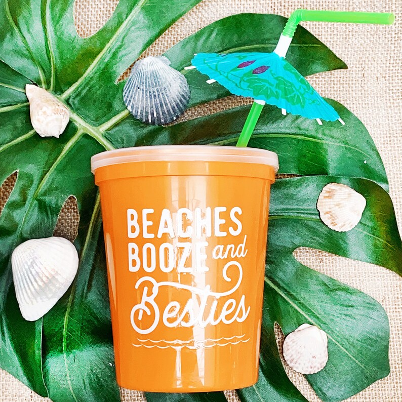 May include: Orange plastic cup with the text "Beaches Booze and Besties" printed on it. A green straw with a blue and green umbrella is sticking out of the cup. The cup is sitting on a green leaf with seashells around it.