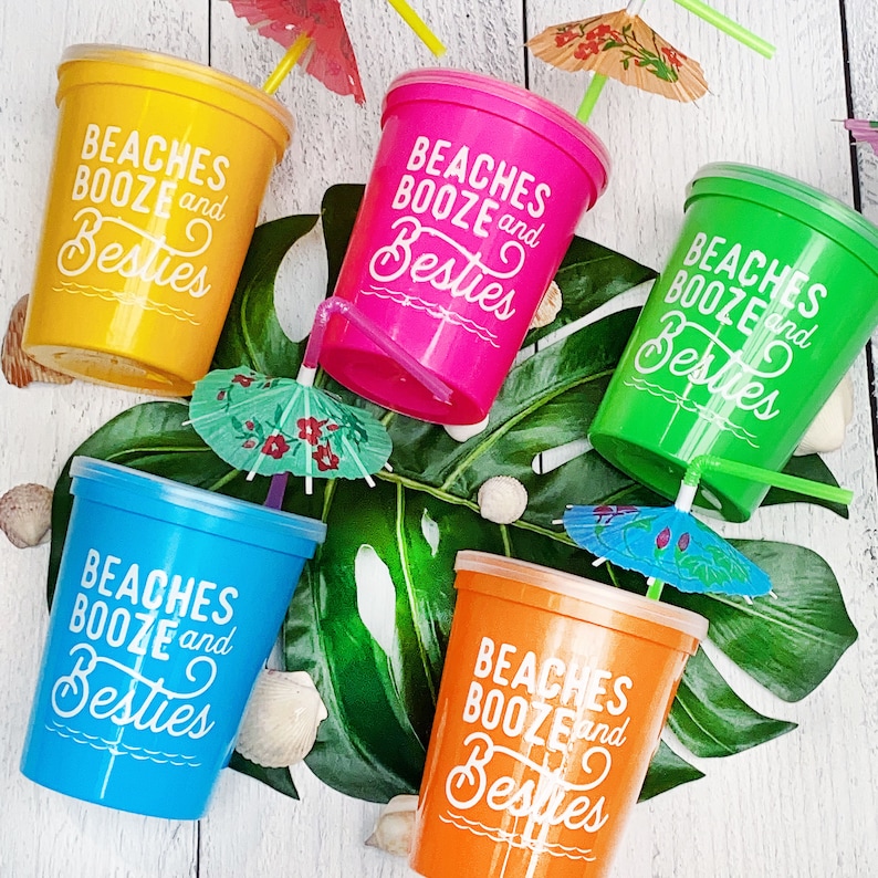 May include: Five colorful plastic cups with lids and straws. Each cup has the text "Beaches Booze and Besties" printed on it. The cups are decorated with small paper umbrellas.