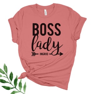 May include: A pink t-shirt with the text "BOSS lady" in black lettering. An arrow with the text "INGRID" is below the text.