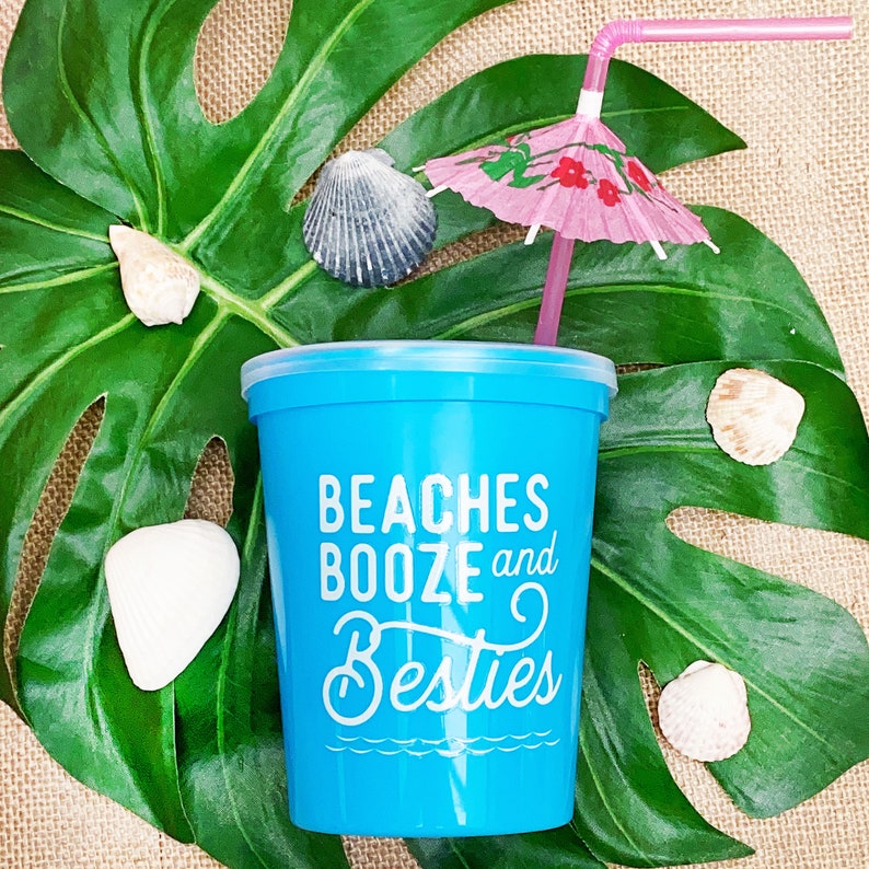 May include: A blue plastic cup with the text "Beaches Booze and Besties" printed on it. The cup is sitting on a green leaf with a pink umbrella and a straw sticking out of it.
