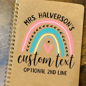 May include: A brown spiral notebook with a gold coil binding. The cover features a rainbow design with a pink, blue, and yellow color scheme. The text "MRS. HALVERSON'S" is printed in black at the top of the cover. The text "custom text" is printed in black below the rainbow. The text "OPTIONAL 2ND LINE" is printed in black below the text "custom text".