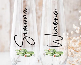May include: Two stemless clear glass champagne flutes featuring a vertical black script name on each glass. The left glass displays the name Simona and the right glass displays the name Winona. Each glass features a green cactus graphic with small orange flowers.