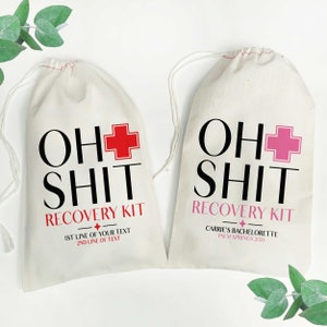 May include: Two off-white drawstring pouches made of fabric. The left bag features black text reading OH SHIT, a red medical cross, and red text reading RECOVERY KIT. The right bag has black text reading OH SHIT, a pink cross, and pink text reading RECOVERY KIT.