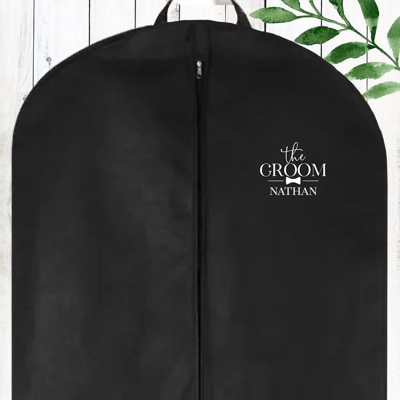 Puede incluir: Bolsa de ropa negra con cierre de cremallera. La bolsa tiene texto blanco que dice "the GROOM NATHAN" con un icono de corbata de lazo.
