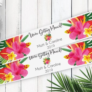 Puede incluir: Dos etiquetas blancas con dise&ntilde;os florales tropicales y el texto "We're Getting Maui'd!" con los nombres Matt & Caroline y el a&ntilde;o 2019.