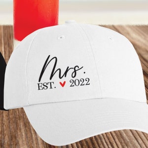 May include: White baseball cap with black text that reads "Mrs. EST. &hearts; 2022".