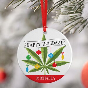 May include: A white round Christmas ornament with a green marijuana leaf design. The ornament has red, green, blue, and yellow ornaments hanging from the leaf. The text "Happy Holidaze" is written in a banner above the leaf. The name "Michaela" is written in red at the bottom of the ornament.