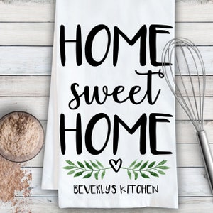 May include: White kitchen towel with the text "Home sweet home" in black cursive font. A small heart and green watercolor leaves are below the text. The text "Beverly's Kitchen" is at the bottom of the towel.