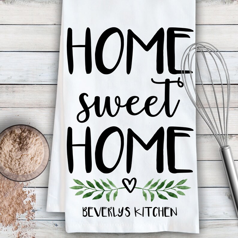 May include: White kitchen towel with the text "Home sweet home" in black cursive font. A small heart and green watercolor leaves are below the text. The text "Beverly's Kitchen" is at the bottom of the towel.