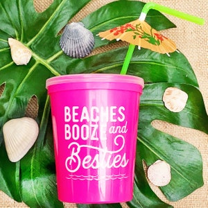 May include: Pink plastic cup with a white design that says "Beaches Booze and Besties". A green straw with a small paper umbrella is in the cup. The cup is sitting on a green leaf.