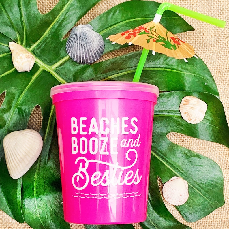 May include: Pink plastic cup with a white design that says "Beaches Booze and Besties". A green straw with a small paper umbrella is in the cup. The cup is sitting on a green leaf.