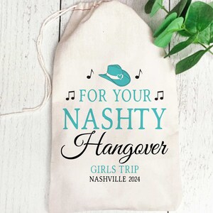 Puede incluir: Una bolsa de lona beige con cord&oacute;n que presenta un icono de sombrero de vaquero turquesa con un coraz&oacute;n, notas musicales negras y el texto FOR YOUR NASHTY Hangover GIRLS TRIP NASHVILLE 2024 impreso en fuentes turquesa y negras en el frente.