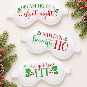 Könnte beinhalten: Drei weiße Schlafmasken aus Satin mit Gummibändern. Jede Maske hat einen anderen weihnachtlichen Spruch in Grün und Rot: "Dreaming of a silent night", "Santa's favorite HO" und "Shhh... I got too LIT."