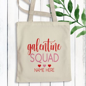 May include: A beige tote bag with the text "galentine SQUAD" in red and pink lettering. Below the text are three red hearts and the text "NAME HERE" in pink.