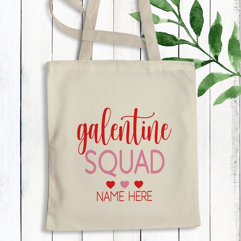 May include: A beige tote bag with the text "galentine SQUAD" in red and pink lettering. Below the text are three red hearts and the text "NAME HERE" in pink.