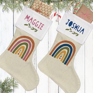 May include: Two beige fabric Christmas stockings featuring a white cuff. The left stocking has the name Maggie in pink with a rainbow graphic. The right stocking has the name Joshua in blue with a rainbow graphic. Each includes a gift box and candy cane.