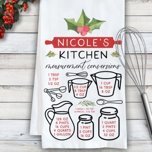 May include: A white kitchen towel with a red and green holly design. The towel features a handwritten inscription "Nicole's Kitchen measurement conversions" and a set of illustrations showing common kitchen measurements in cups, ounces, tablespoons, teaspoons, pints, quarts, and gallons.