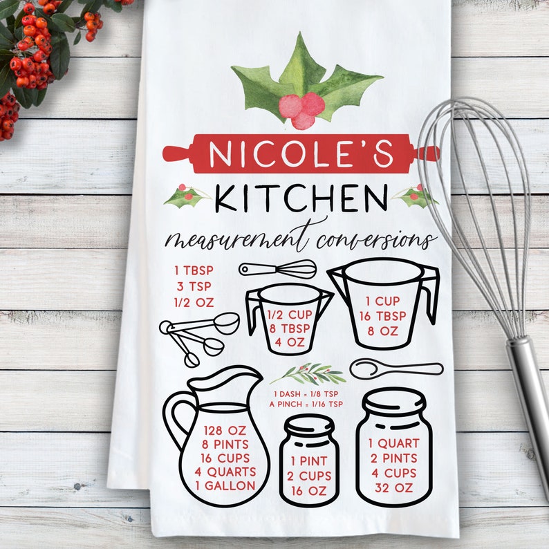 May include: A white kitchen towel with a red and green holly design. The towel features a handwritten inscription "Nicole's Kitchen measurement conversions" and a set of illustrations showing common kitchen measurements in cups, ounces, tablespoons, teaspoons, pints, quarts, and gallons.