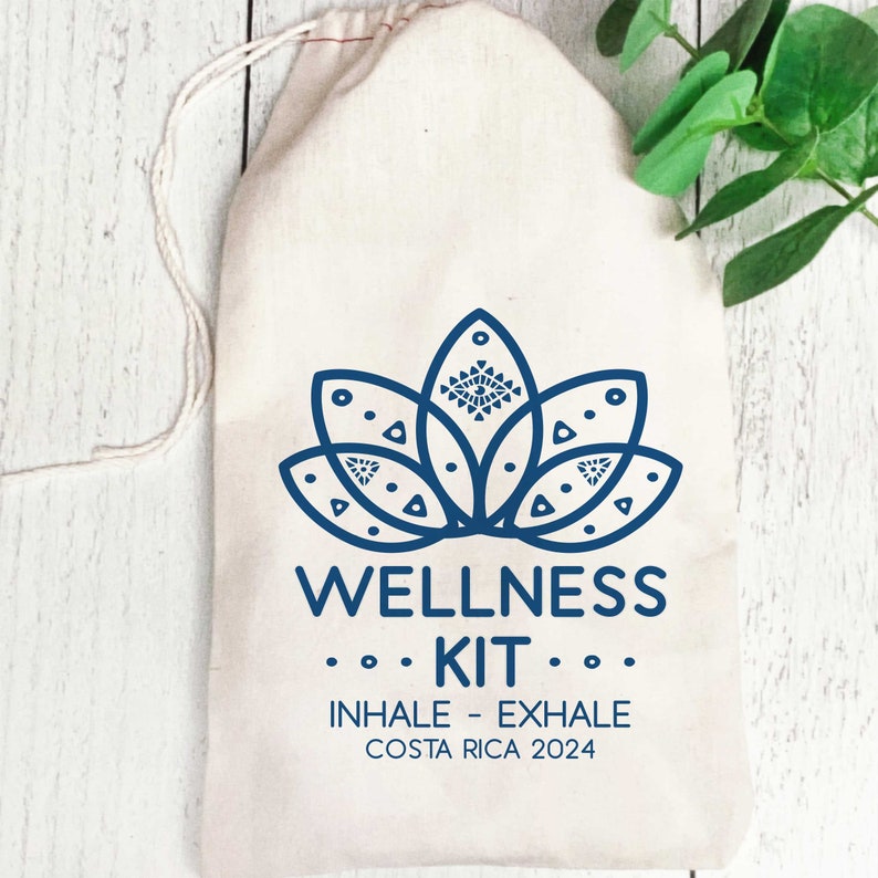 K&ouml;nnte beinhalten: Ein beigefarbener Baumwollbeutel mit Kordelzug, der mit einer dunkelblauen Grafik einer Lotusblume mit geometrischen Mustern bedruckt ist. Darunter steht der Text WELLNESS KIT, INHALE - EXHALE, COSTA RICA 2024 in einer klaren Schrift.