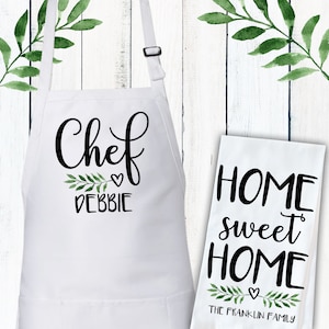 May include: White kitchen apron with black text that says "Chef Debbie" with a small heart and green leaves. A white tea towel with black text that says "Home sweet Home" with a small heart and green leaves. The text below the leaves says "The Franklin Family".