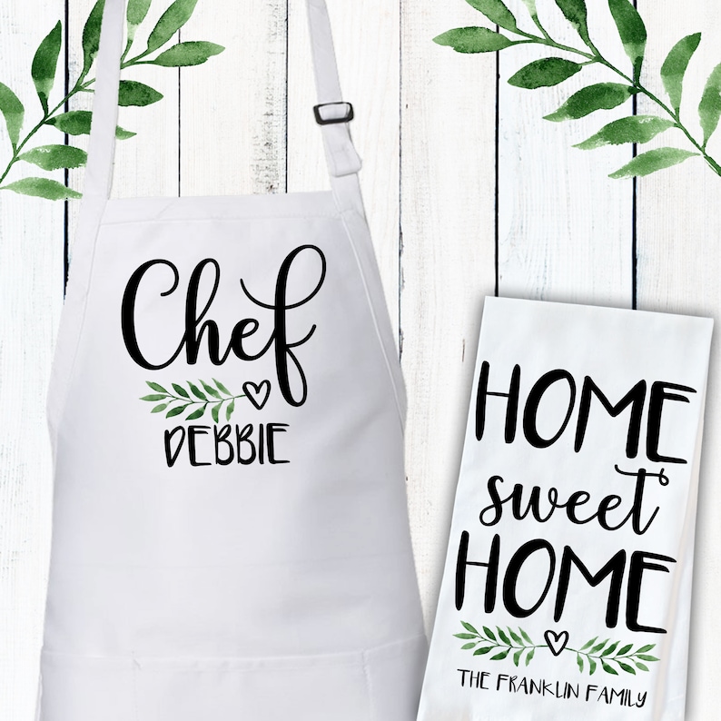 May include: White kitchen apron with black text that says "Chef Debbie" with a small heart and green leaves. A white tea towel with black text that says "Home sweet Home" with a small heart and green leaves. The text below the leaves says "The Franklin Family".