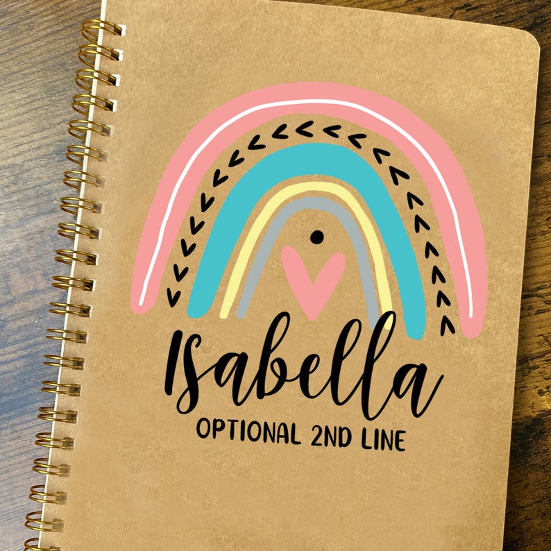 May include: A brown spiral notebook with a rainbow design and the name "Isabella" written in black cursive. The rainbow is made up of pink, blue, yellow, and gray arcs with a pink heart in the center. The text "OPTIONAL 2ND LINE" is written below the name.
