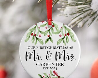 May include: White ceramic Christmas ornament with a red ribbon. The ornament features a watercolor design of green leaves and red berries with the text "Our First Christmas as Mr. & Mrs. CARPENTER EST. 2024".