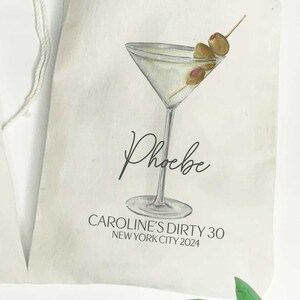Może przedstawiać: Biała karta z ilustracją kieliszka do martini i zielonych oliwek. Tekst "Phoebe" jest napisany kursywą pod kieliszkiem. Tekst "CAROLINE'S DIRTY 30 NEW YORK CITY 2024" jest wydrukowany pod nazwą.