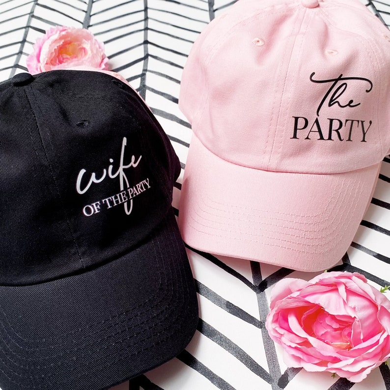 May include: Two baseball caps, one black and one pink. The black cap says "Wife of the Party" in white script. The pink cap says "The Party" in black script.