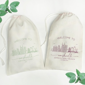 May include: Two off-white drawstring bags with a Nashville skyline design. One bag has green text that reads "WELCOME TO nashville" with the names "CAROLINE & CASEY" and the year "2026". The other bag has burgundy text that reads "WELCOME TO nashville" with "JOHNSON FAMILY REUNION OCTOBER 2026".
