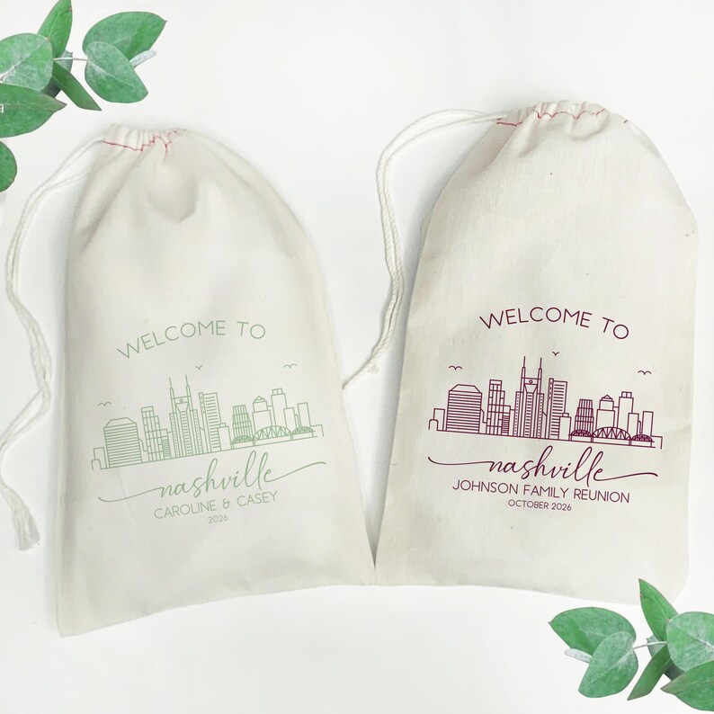 May include: Two off-white drawstring bags with a Nashville skyline design. One bag has green text that reads "WELCOME TO nashville" with the names "CAROLINE & CASEY" and the year "2026". The other bag has burgundy text that reads "WELCOME TO nashville" with "JOHNSON FAMILY REUNION OCTOBER 2026".