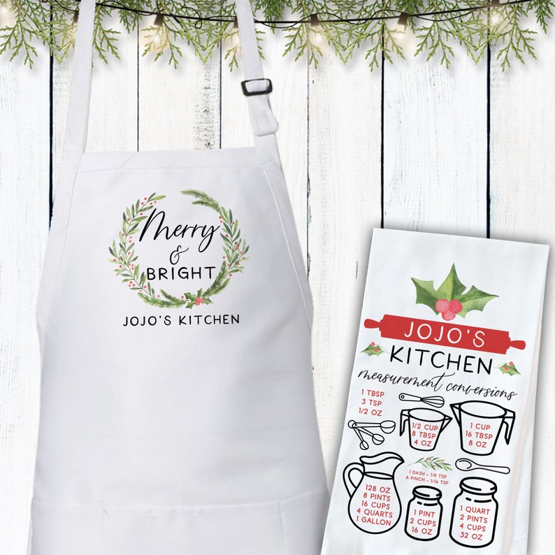 May include: White kitchen apron with a green wreath and the text "Merry & Bright" and "JOJO'S KITCHEN". A white tea towel with a red holly design and the text "JOJO'S KITCHEN measurement conversions" with a list of common kitchen measurements.