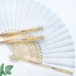 May include: White paper fan with a pleated design, partially open, with three wooden fans. The wooden fans have the text "LAUREN'S NOLARETTE" and "BEIGNETS, BUBBLES & BASTILLE +++" printed on them. The fans are a light beige color.