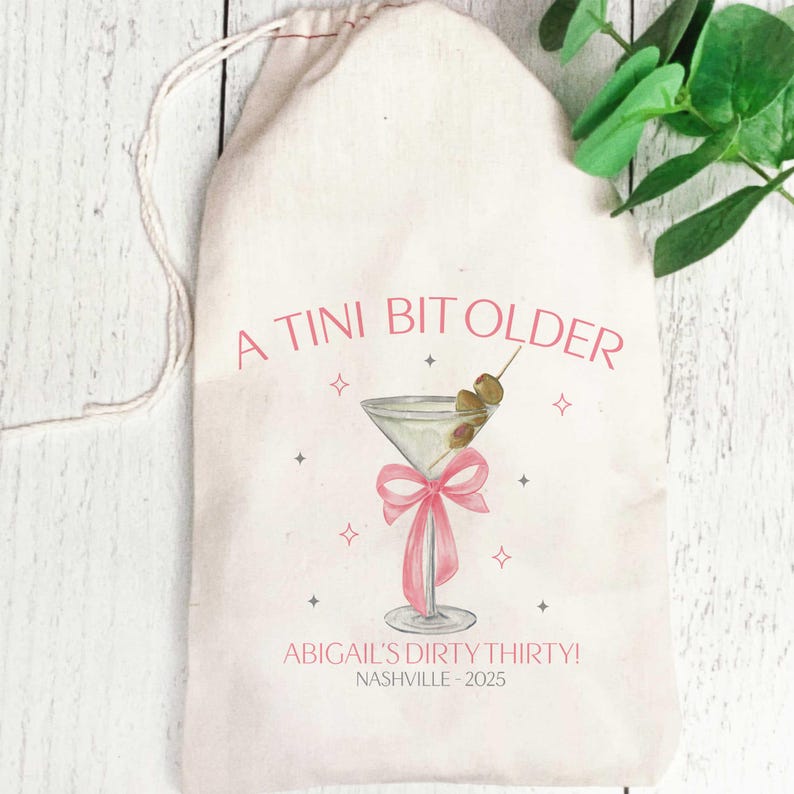 May include: A white canvas bag with a pink and grey illustration of a martini glass with olives and a pink bow. The text "A TINI BIT OLDER" is printed in pink on the bag. The text "ABIGAIL'S DIRTY THIRTY! NASHVILLE - 2025" is printed in grey at the bottom of the bag.