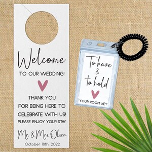 May include: Welcome to our wedding door hanger with a pink heart and the text "Welcome to our wedding! Thank you for being here to celebrate with us! Please enjoy your stay. Mr. & Mrs. Olsen October 18th, 2022". A clear plastic room key holder with a black elastic band attached to it. The holder has the text "To have & to hold" with a pink heart and the text "Your room key".