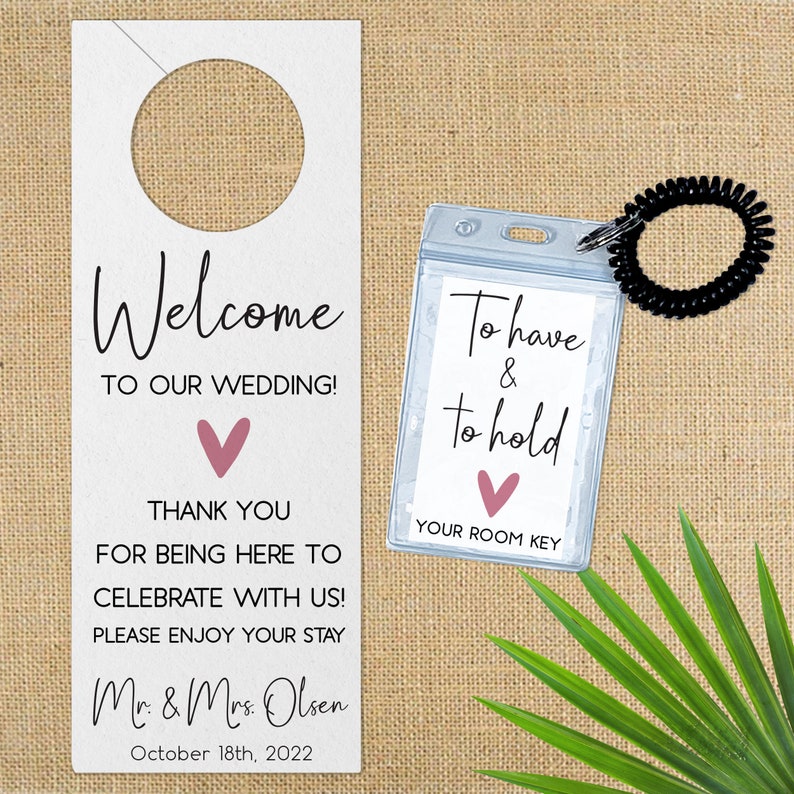 May include: Welcome to our wedding door hanger with a pink heart and the text "Welcome to our wedding! Thank you for being here to celebrate with us! Please enjoy your stay. Mr. & Mrs. Olsen October 18th, 2022". A clear plastic room key holder with a black elastic band attached to it. The holder has the text "To have & to hold" with a pink heart and the text "Your room key".