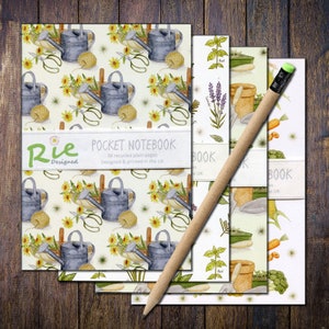 May include: A collection of pocket notebooks featuring garden-themed designs, such as watering cans, flowers, and vegetables. The notebooks have a white band with the text "Pocket Notebook" and a pencil with a green eraser rests on top. The notebooks are on a wooden surface.